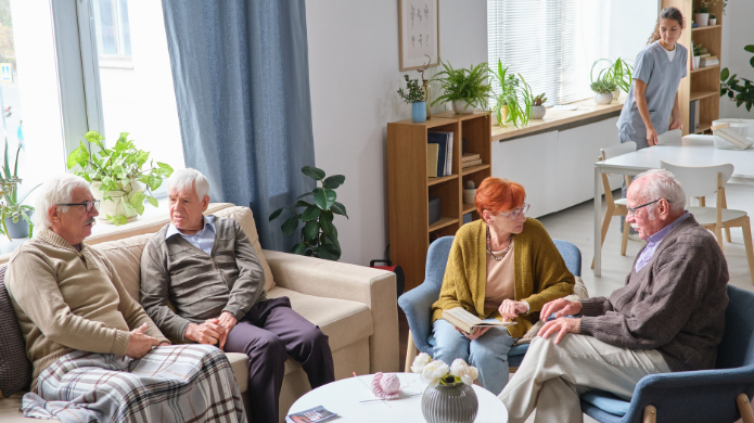 Betreutes Wohnen Vier ältere Menschen sitzen in einem hellen Gemeinschaftsraum zusammen und unterhalten sich. Im Hintergrund steht eine jüngere Frau an einem Esstisch.