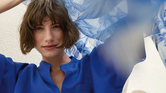 Time to Shine Nahaufnahme einer lächelnden Frau in einem kräftig blauen Oberteil, die ein blau-weiß gemustertes Tuch hochhält.