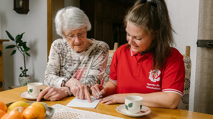 Eine Haushaltshilfe für Senioren plant gemeinsam mit einer Seniorin am Tisch den nächsten Einkauf.