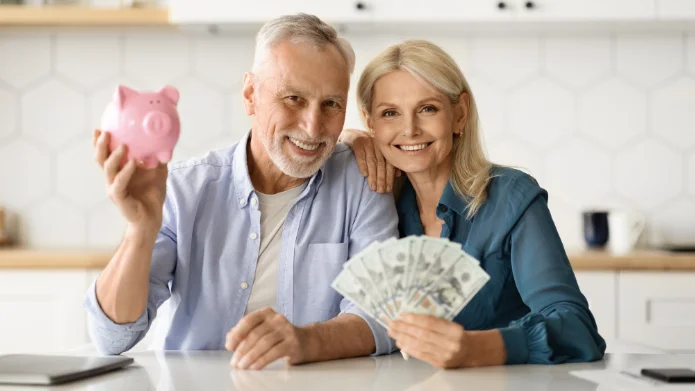 Steuern sparen als Rentner Älteres Paar sitzt am Tisch und hält ein Sparschwein sowie Bargeld in der Hand.