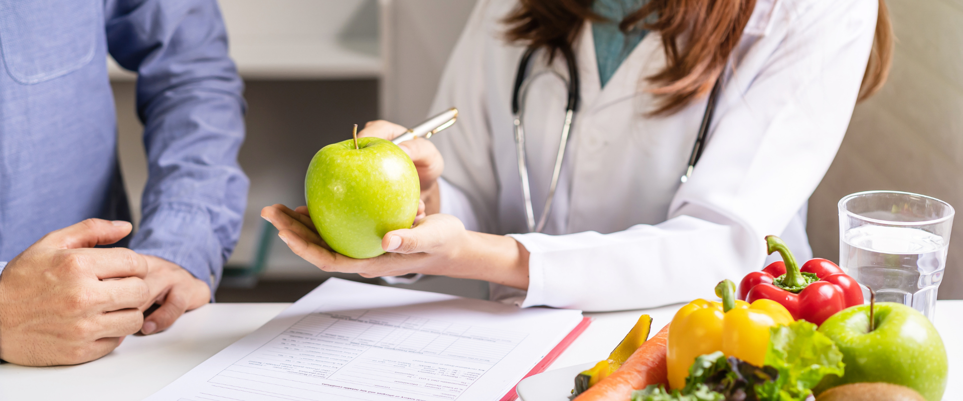 Ernährungsberatung Eine Ernährungsberaterin zeigt einer Person einen grünen Apfel; auf dem Tisch liegen frisches Obst und Gemüse.