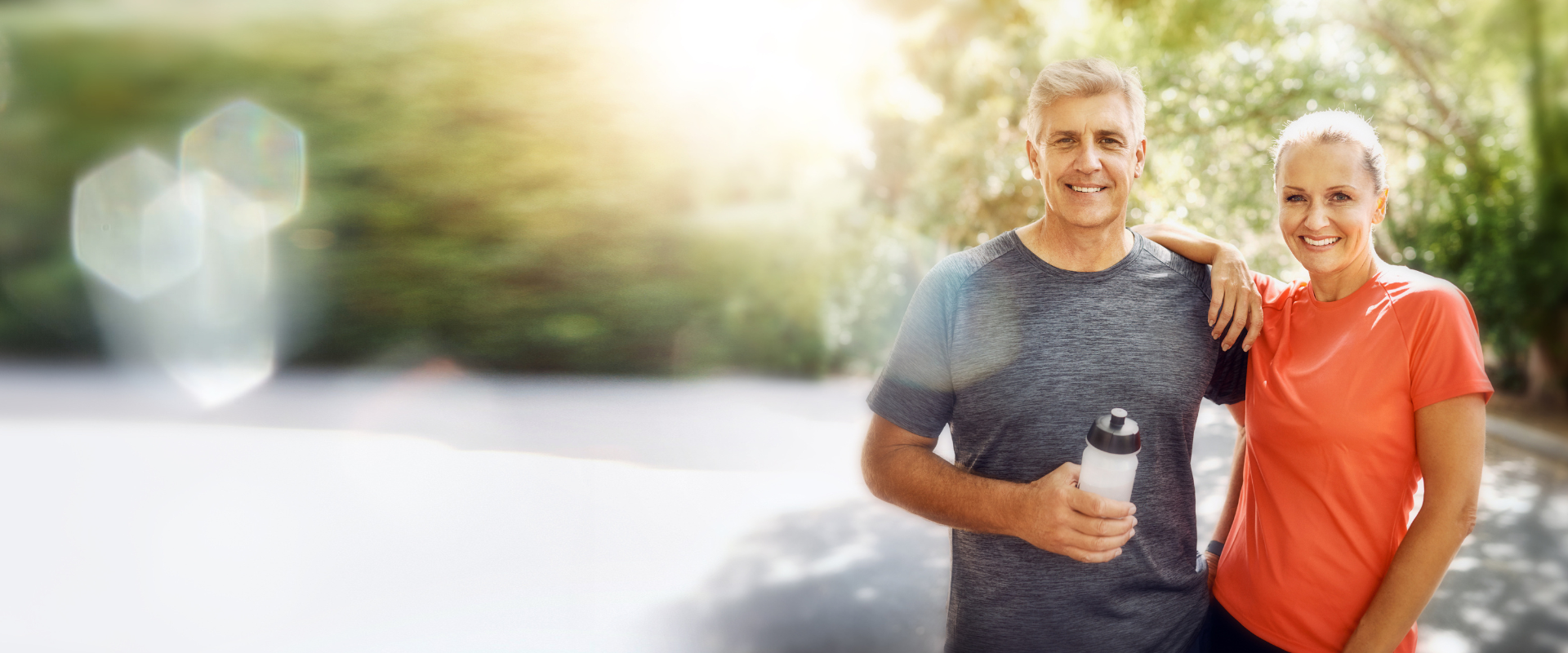 Fitnesstraining Ein sportlich gekleidetes Paar steht nach dem Training im Freien und lächelt in die Kamera; der Mann hält eine Trinkflasche in der Hand.
