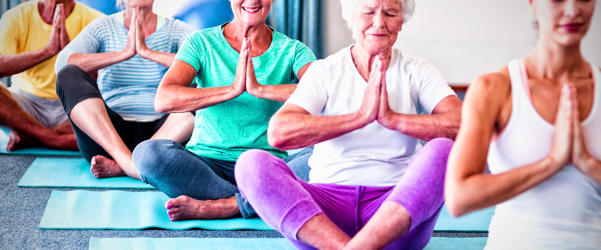 Yoga Mehrere ältere Menschen sitzen in einem Yogakurs auf Matten und halten mit gefalteten Händen eine ruhige Sitzposition.