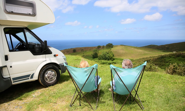 Zwei Menschen sitzen in Campingstühlen neben einem Wohnmobil und genießen den Ausblick auf ein großes Tal. 
