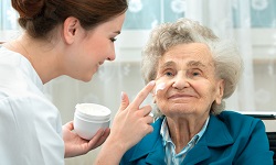 Pflegerin cremt einer älteren Dame das Gesicht ein.